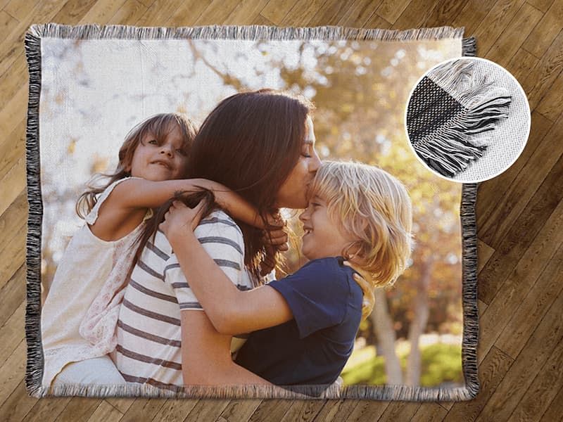A woven fringed photo throw blanket on a wood floor, showing a mother kissing her two young children, with a close-up inset of the fringe detail.