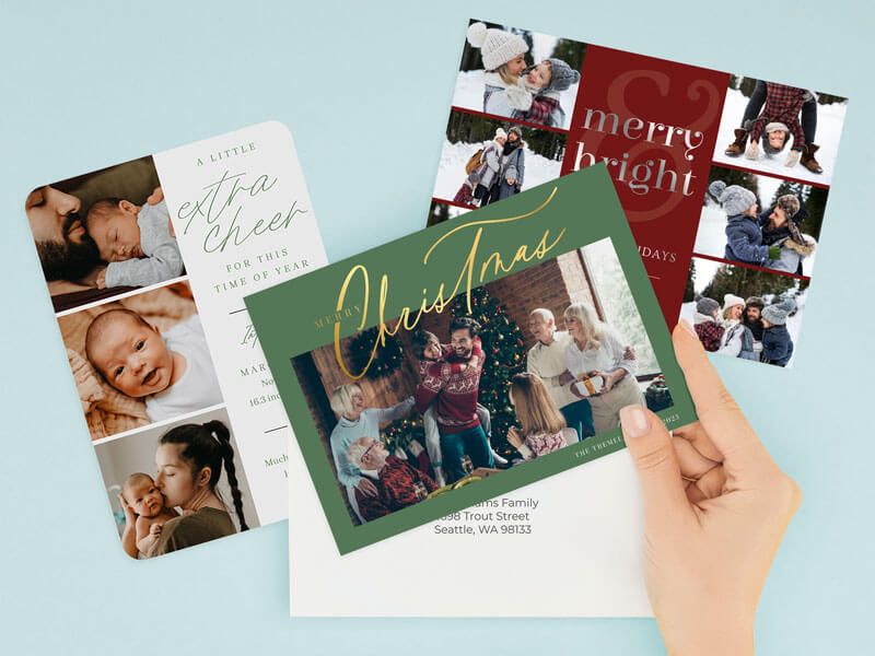 A hand holding a green 'Merry Christmas' family holiday card over an envelope, with a red 'Merry & Bright' snow-scene card and a white 'A Little Extra Cheer' baby announcement card layered behind it.