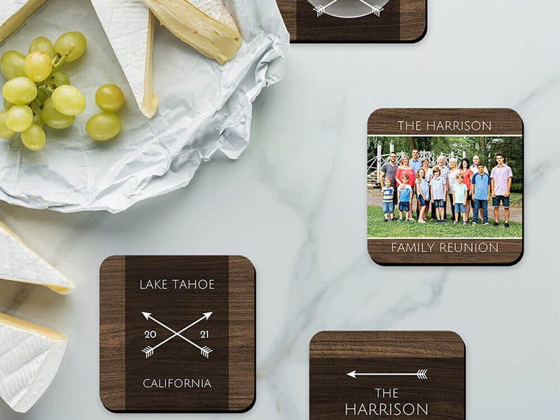 Four wood-look photo coasters on a marble counter, one printed with a family reunion photo and others personalized with 'Lake Tahoe 2021 California' and 'The Harrison' text, near a cheese board with grapes.