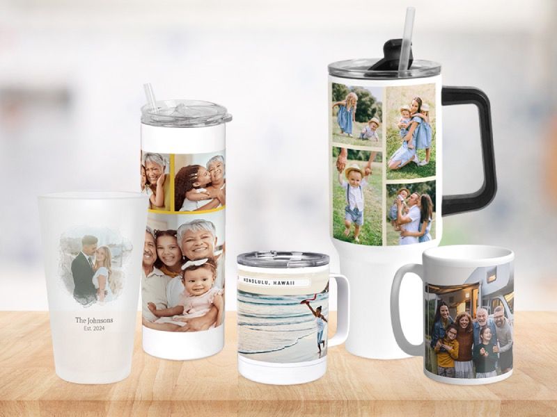 Five personalized photo drinkware pieces on a wooden table: a frosted pint glass, a tall tumbler with straw, a handled travel mug, a large handled tumbler, and a coffee mug, each printed with different family and travel photos.
