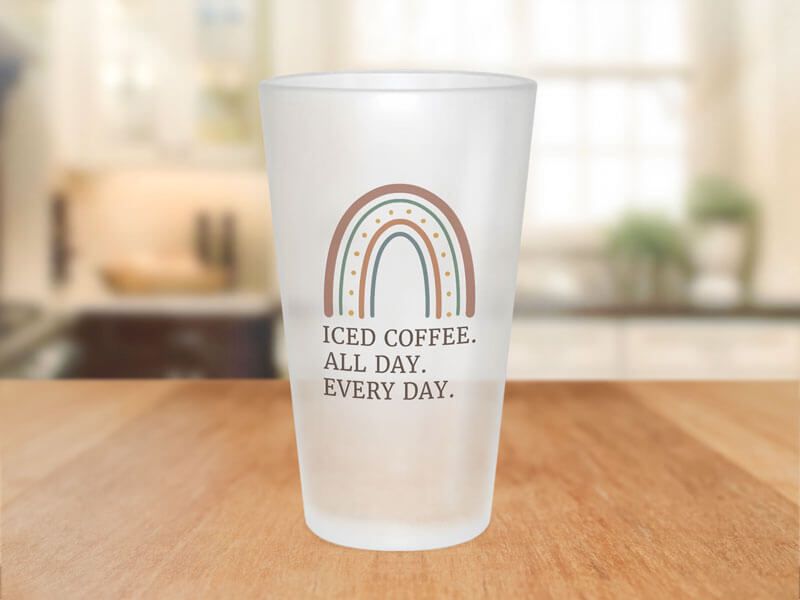 A frosted glass tumbler on a wooden table printed with a boho rainbow design and the text 'Iced Coffee. All Day. Every Day.'