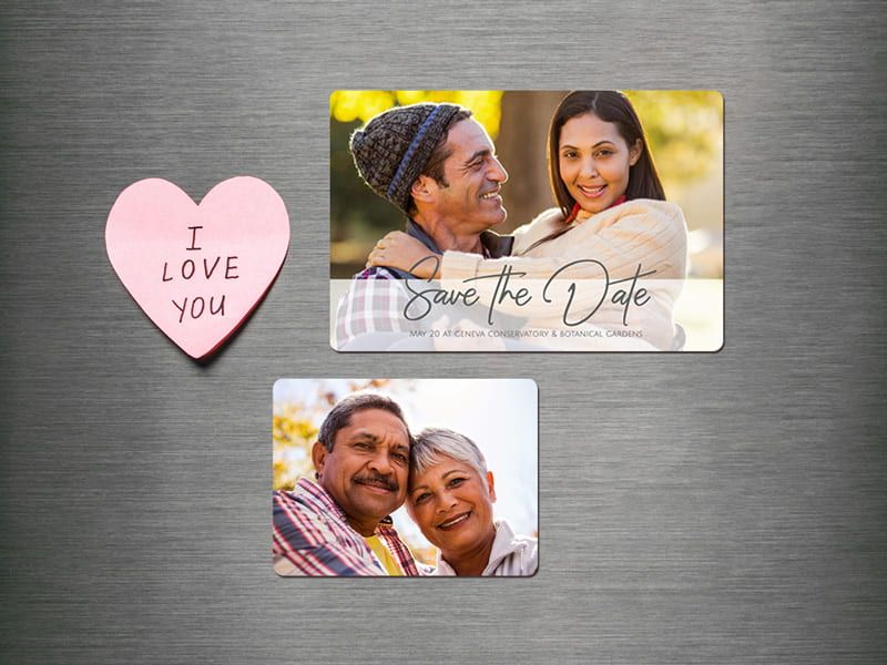 Two photo magnets on a metal refrigerator: a 'Save the Date' magnet of a couple embracing outdoors, and a plain photo magnet of an older couple, next to a pink heart note reading 'I Love You.'