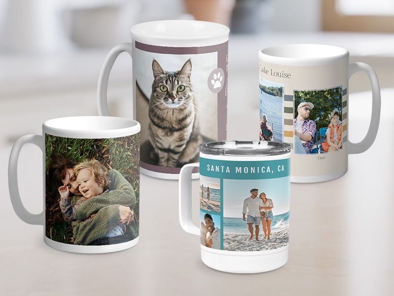 Four personalized photo mugs on a counter: one with a mother and child in grass, one with a tabby cat portrait, one with 'Lake Louise' family photos, and an insulated travel mug labeled 'Santa Monica, CA' with beach photos.