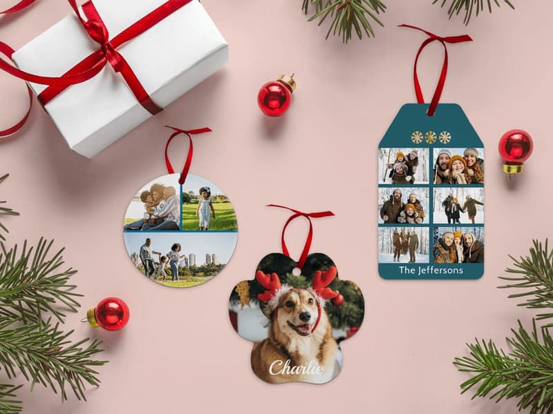 Three Christmas photo ornaments on a pink background with pine branches: a round ornament with family photos, a paw-shaped ornament of a dog wearing antlers labeled 'Charlie,' and a teal tag ornament with winter family photos reading 'The Jeffersons.'