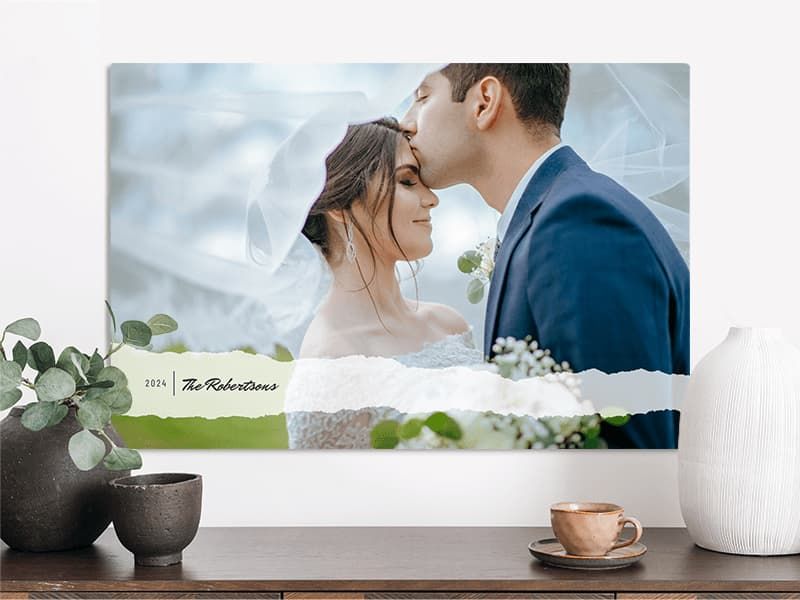 A wide canvas print of a groom kissing a bride's forehead under her veil, labeled '2024 The Robertsons,' hanging above a wood console table with a vase and teacup.