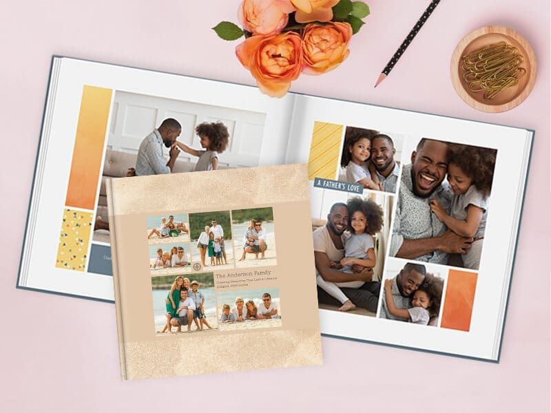An open photo book showing a father-and-daughter photo spread titled 'A Father's Love,' with a closed hardcover 'The Anderson Family' photo book styled with beach photos propped on top, surrounded by flowers, a pencil, and paper clips.