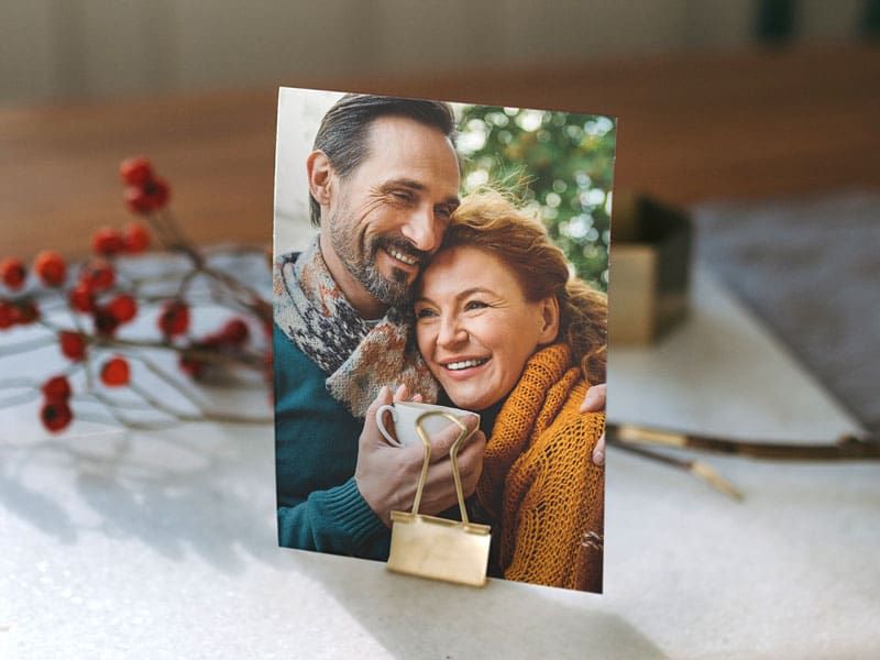 A single photo print held upright by a gold binder clip, showing a smiling couple embracing outdoors while the man holds a mug, displayed on a table with red berry sprigs.