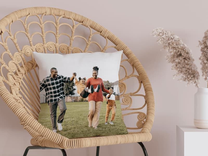 A white throw pillow printed with a photo of a family running on grass, resting on a woven rattan peacock chair.