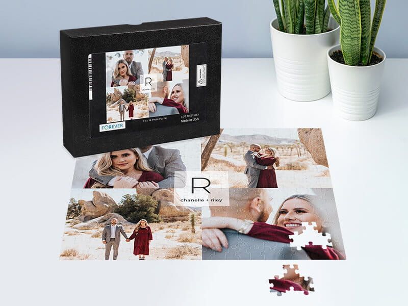 A boxed jigsaw puzzle and its assembled portion on a white desk, featuring engagement photos of a couple in the desert labeled 'chanelle + riley,' with potted plants in the background.