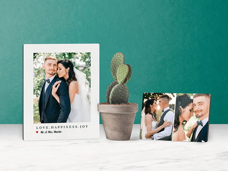 Two tabletop photo panels on a marble surface against a green wall: a framed-style panel of a bride and groom reading 'Love. Happiness. Joy. Mr. & Mrs. Martin,' and a smaller panel with two wedding photos, beside a potted cactus.