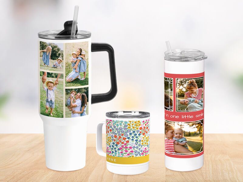 Three personalized drinkware pieces on a wooden surface: a large handled tumbler with a family photo collage, a floral-print mug personalized with a name, and a tall straw tumbler with a red-bordered photo design of a mother and children.