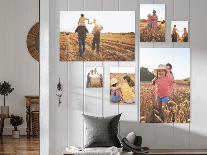 A gallery wall of six canvas photo prints in varying sizes on a white paneled wall, all showing a family in a golden wheat field, arranged above an entryway bench with a pillow, blanket, and hat.